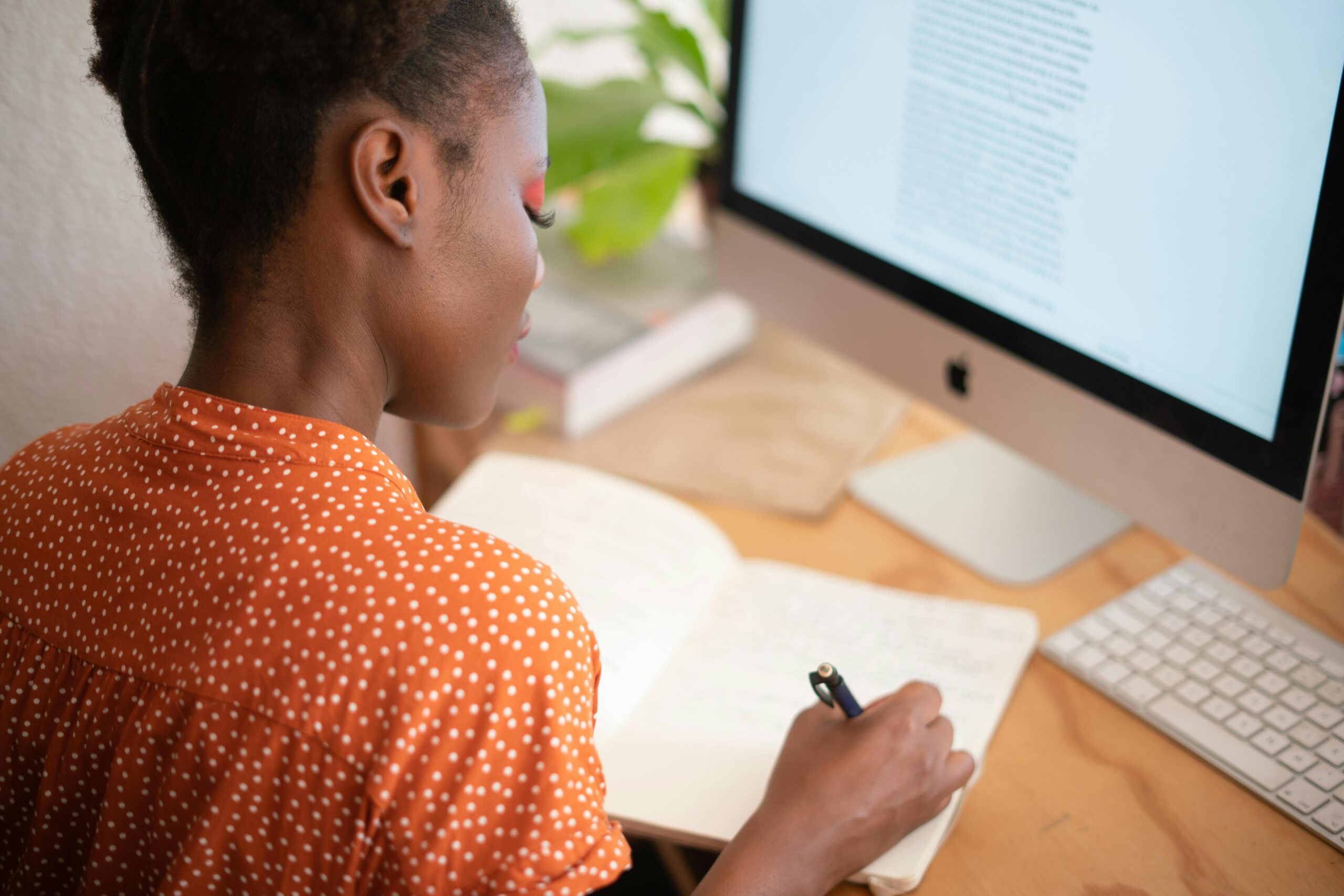 A woman in a polka-dot shirt writes in a notebook at her desk with a computer screen visible.