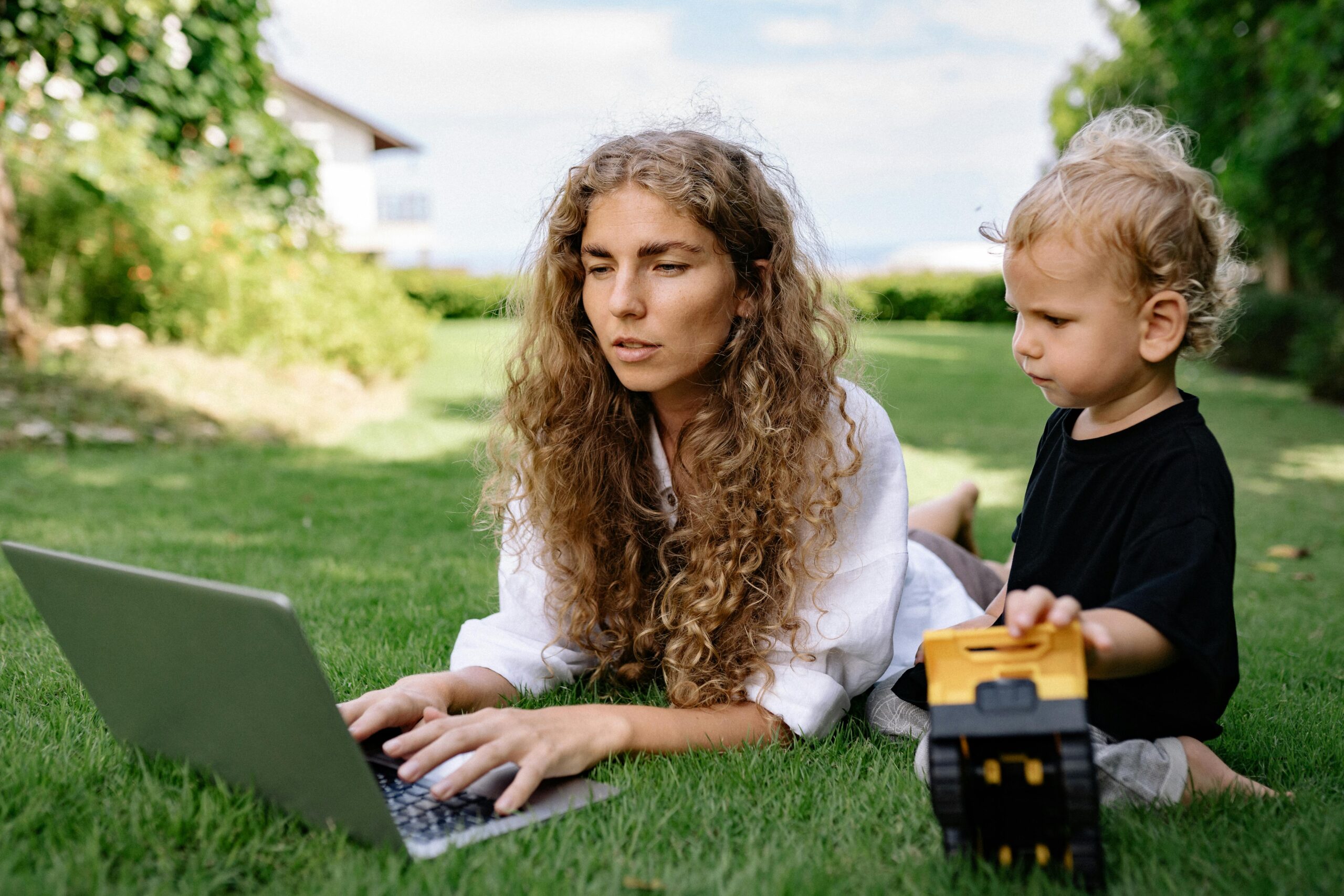 Woman working on a laptop while lying on grass with her child nearby playing.